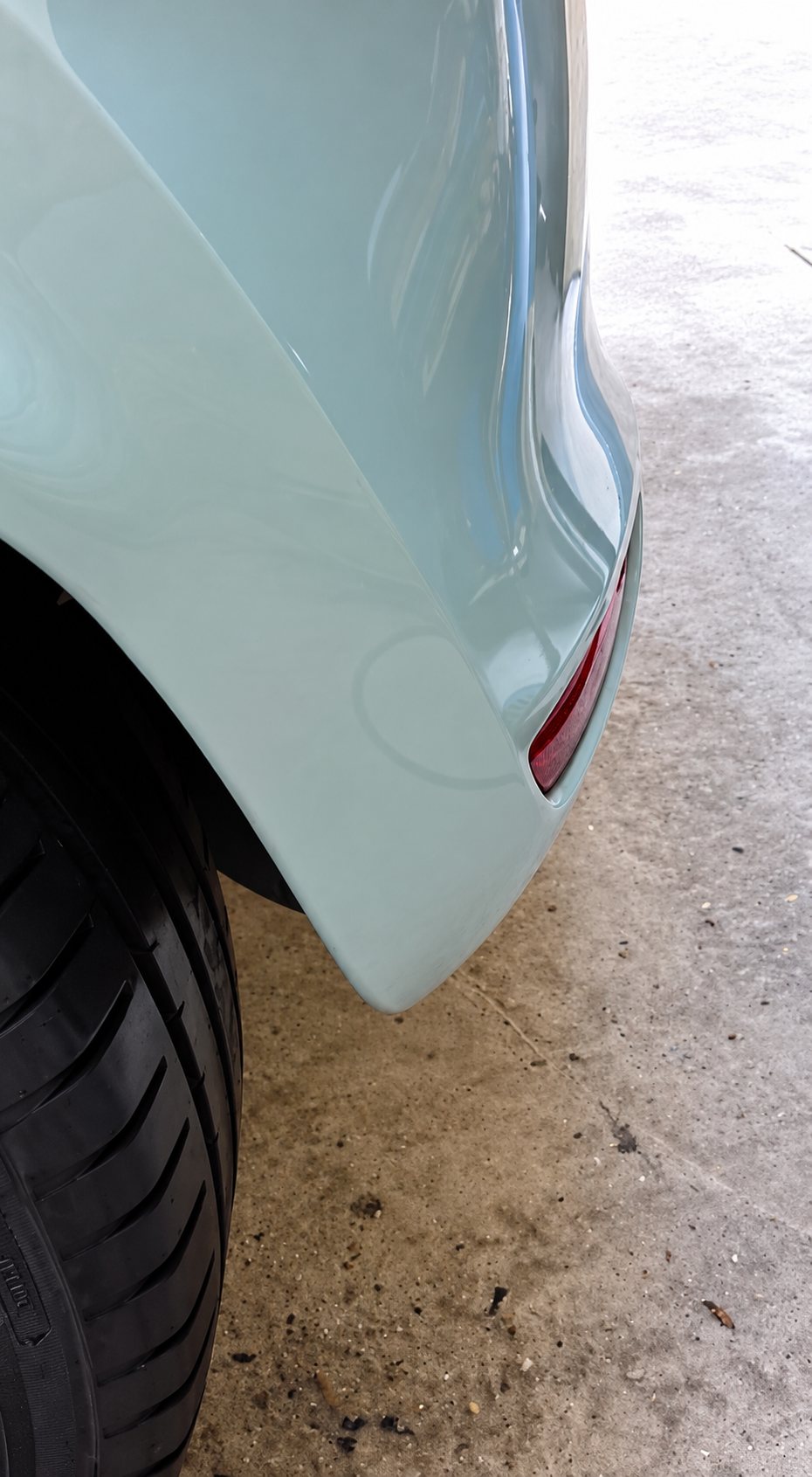 Gros plan sur l'aile arrière et le pare-chocs d'une voiture bleu pâle garée dans un garage, à côté d'un pneu noir.