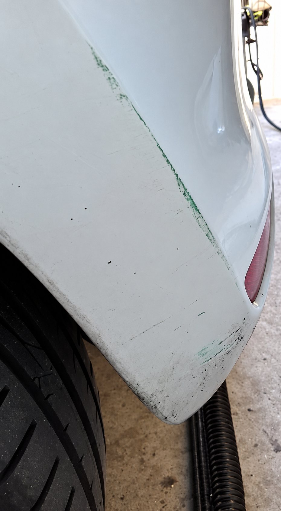 Gros plan sur l'aile arrière d'une voiture vert clair, avec des rayures et des traces de ponçage, au-dessus d'un pneu dans un garage.