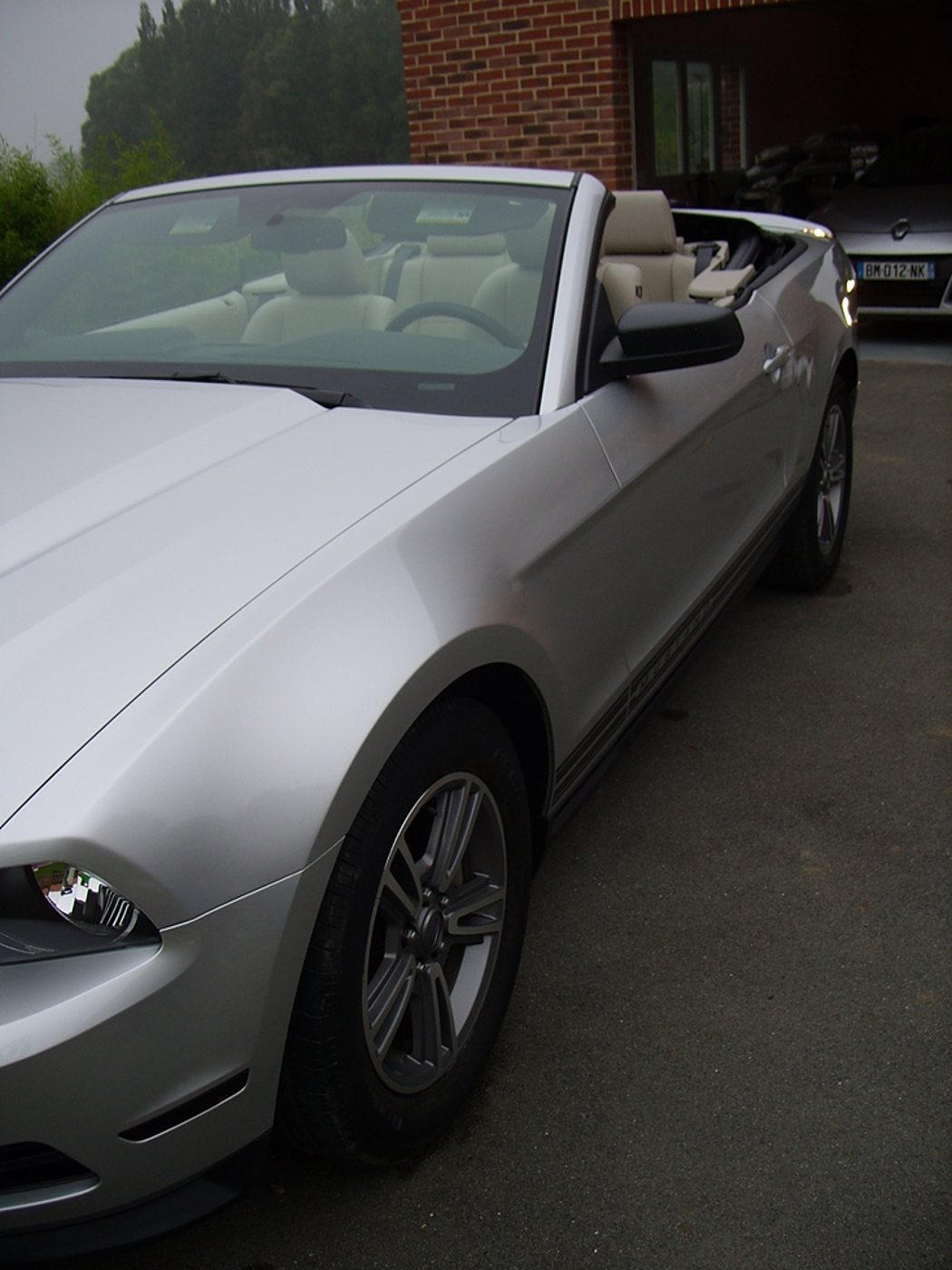 Une Ford Mustang cabriolet grise aux sièges beiges est garée sur une allée devant un bâtiment en briques rouges, avec une autre voiture en arrière-plan.