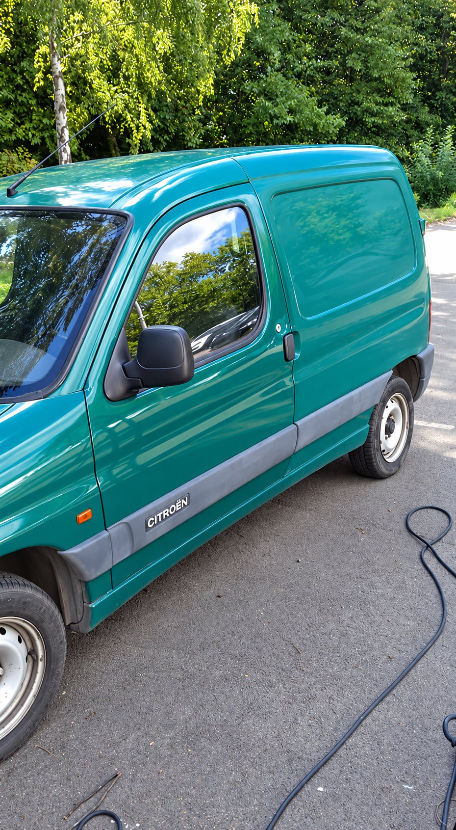 Une camionnette utilitaire Citroën verte est garée sur un sol en bitume devant des arbres, avec un tuyau noir posé à côté.
