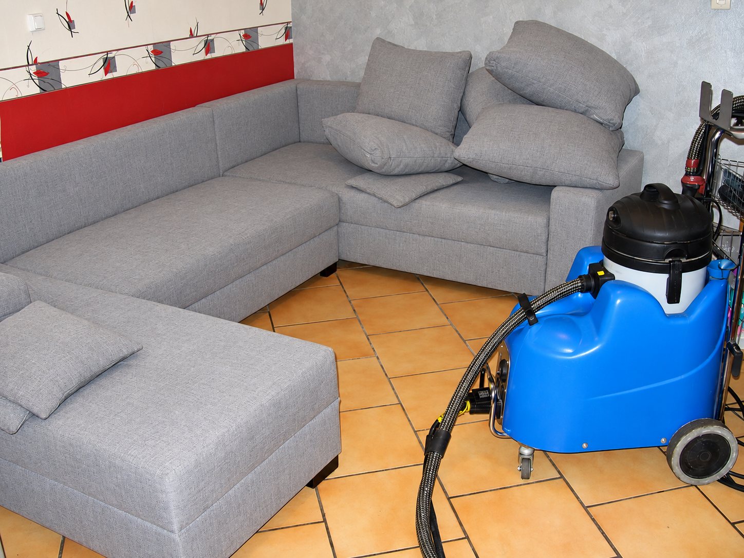 Un canapé d'angle gris dans un salon au carrelage orangé, à côté d'une machine de nettoyage bleue avec son tuyau, utilisée pour le nettoyage des textiles.