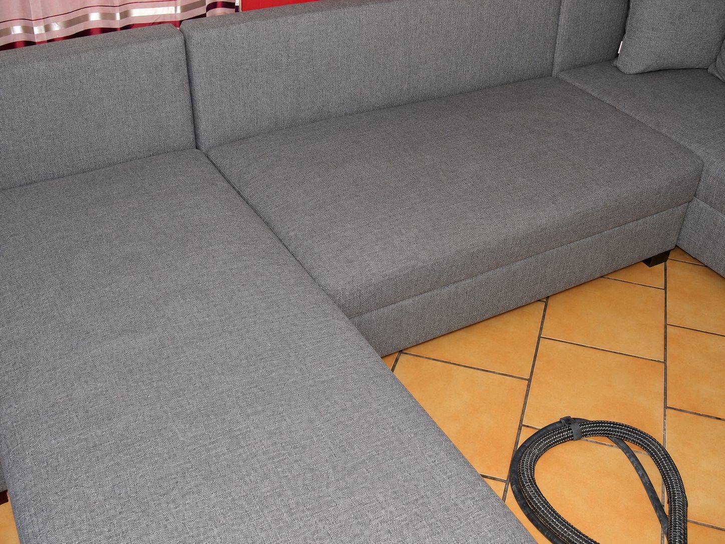 Un canapé d'angle en tissu gris posé sur un carrelage orange, avec le tuyau d'une machine de nettoyage au sol.