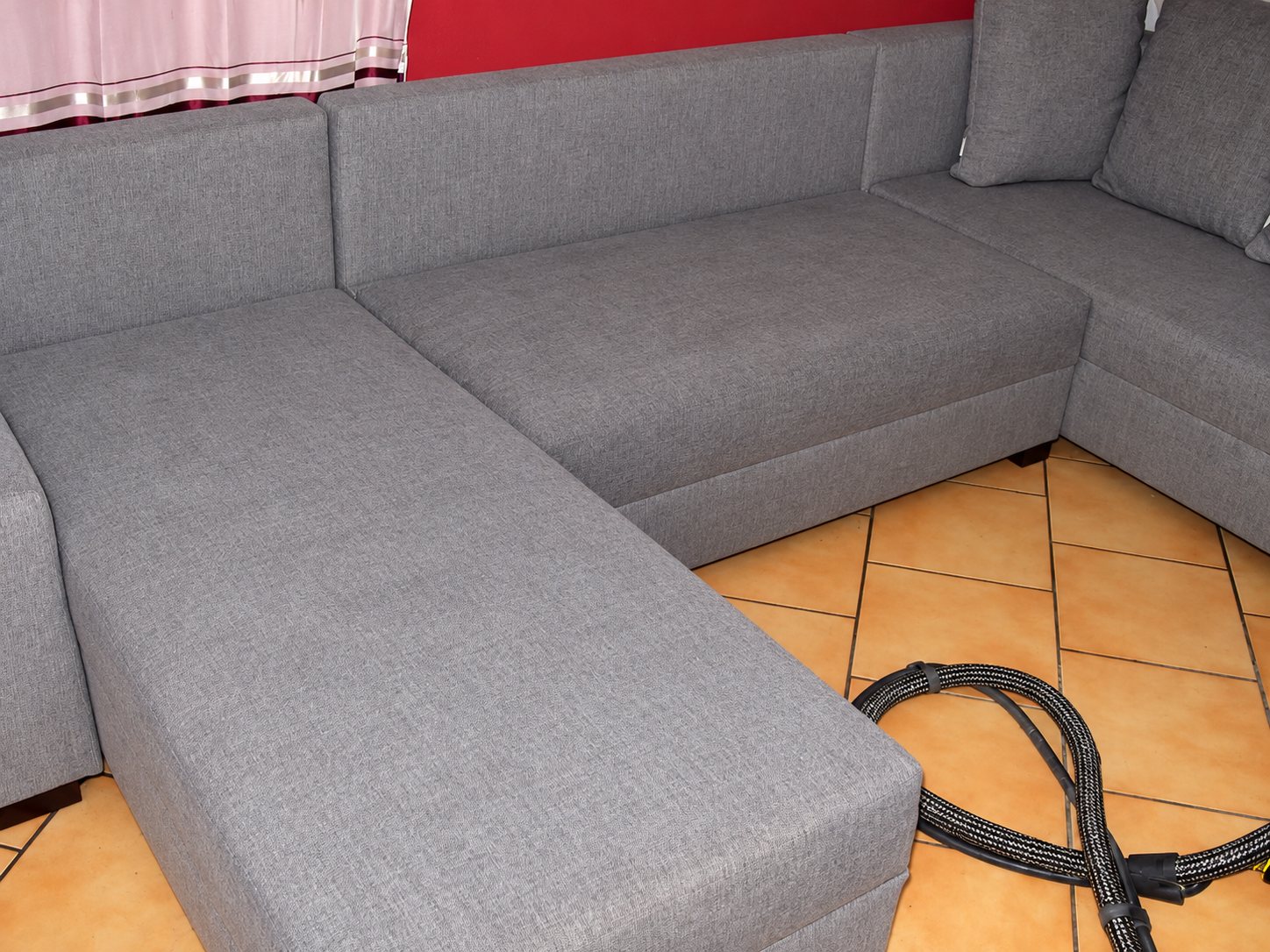 Un grand canapé d'angle en tissu gris, fraîchement nettoyé, posé sur un carrelage orangé avec le tuyau d'une machine de nettoyage au premier plan.