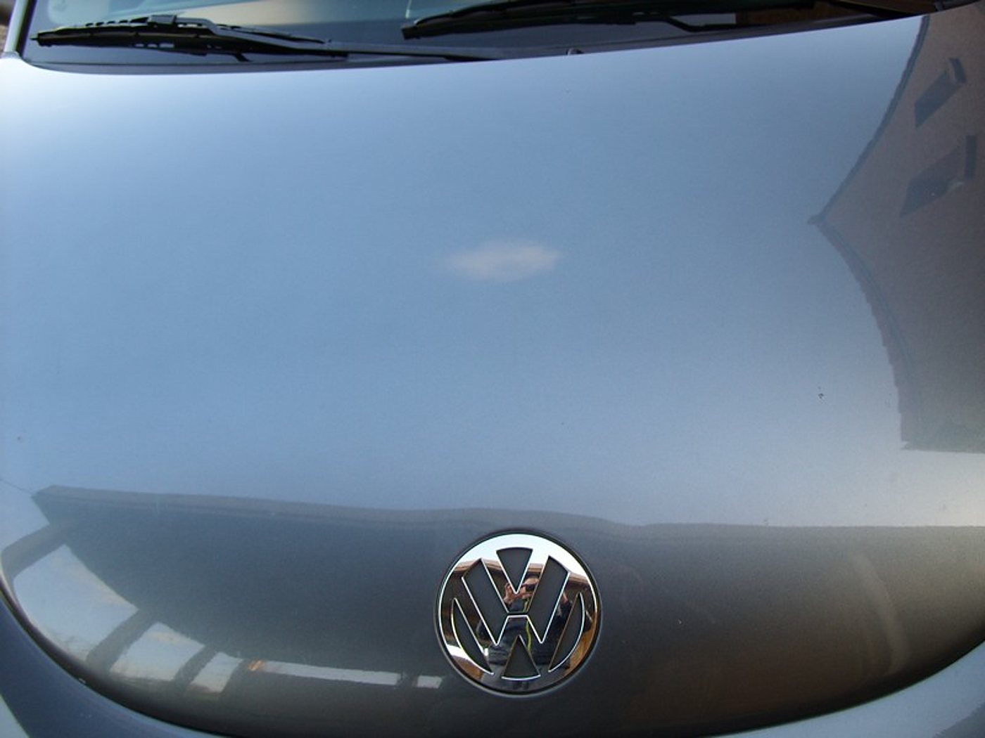 Gros plan sur le capot lisse et brillant d'une voiture grise ornée du logo Volkswagen chromé, avec des reflets de bâtiment.