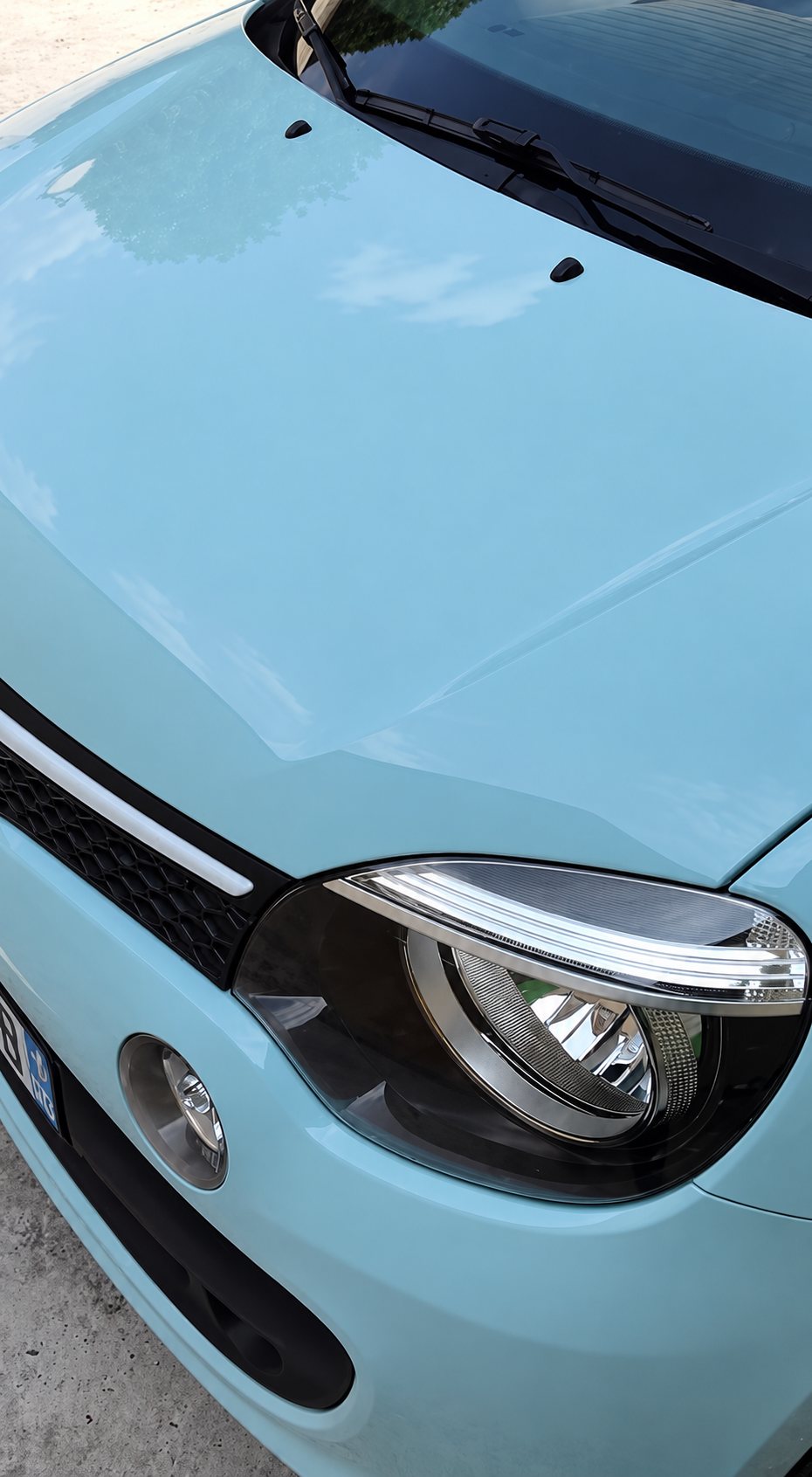 Gros plan sur le capot lisse et le phare avant d'une petite voiture bleu pastel garée sur un sol de gravier clair.
