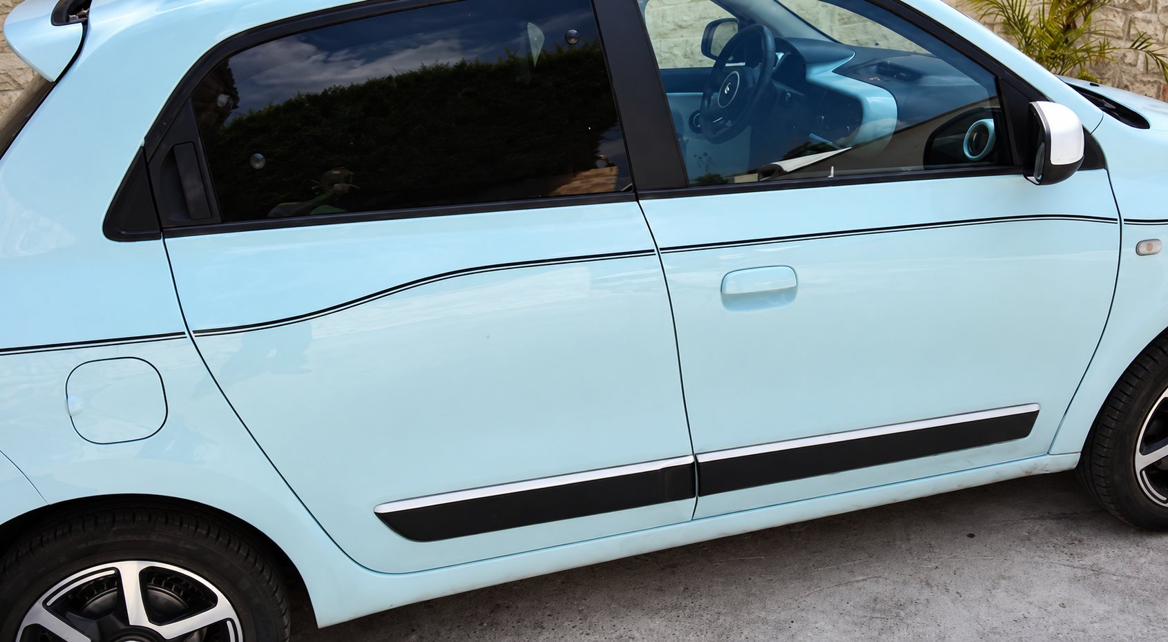 Vue latérale d'une petite voiture bleu pastel à quatre portes garée sur un sol en béton, avec des filets noirs sur la carrosserie.