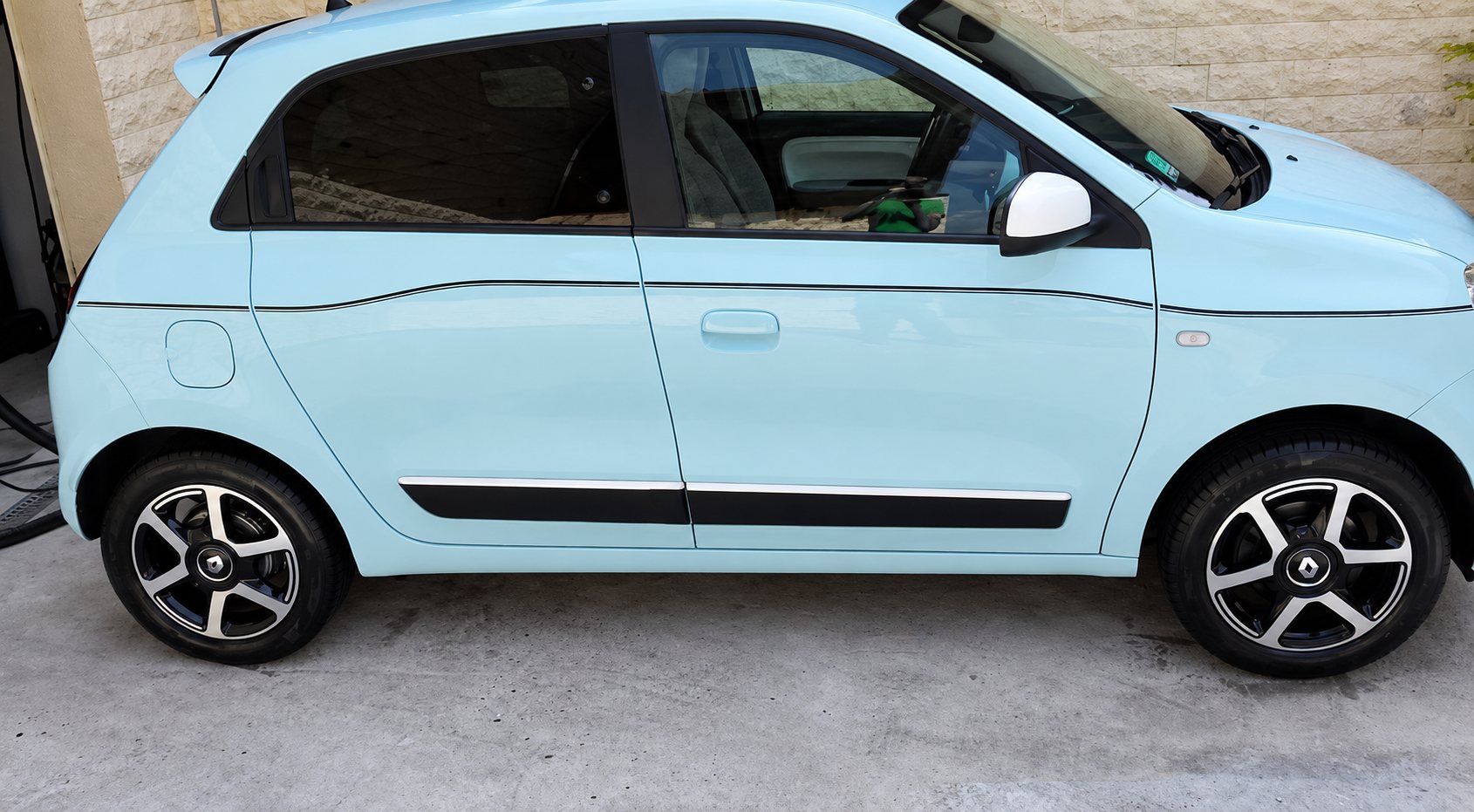 Une petite voiture citadine bleu pastel à quatre portes, photographiée de côté, est garée sur un sol en béton devant un mur en pierre claire.