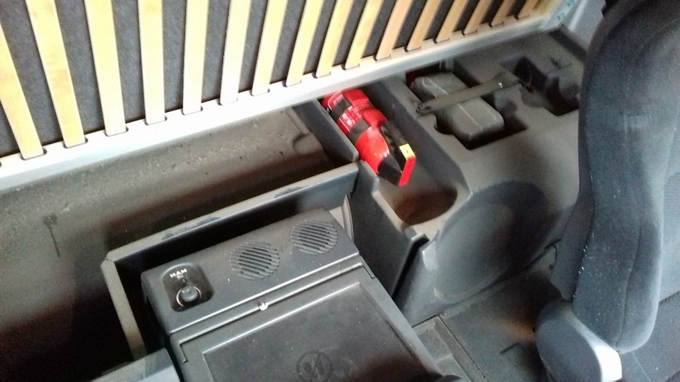 Vue de l'intérieur d'une cabine de camion montrant un compartiment de rangement avec une perceuse-visseuse rouge, un petit réfrigérateur et un siège noir sous un sommier à lattes.
