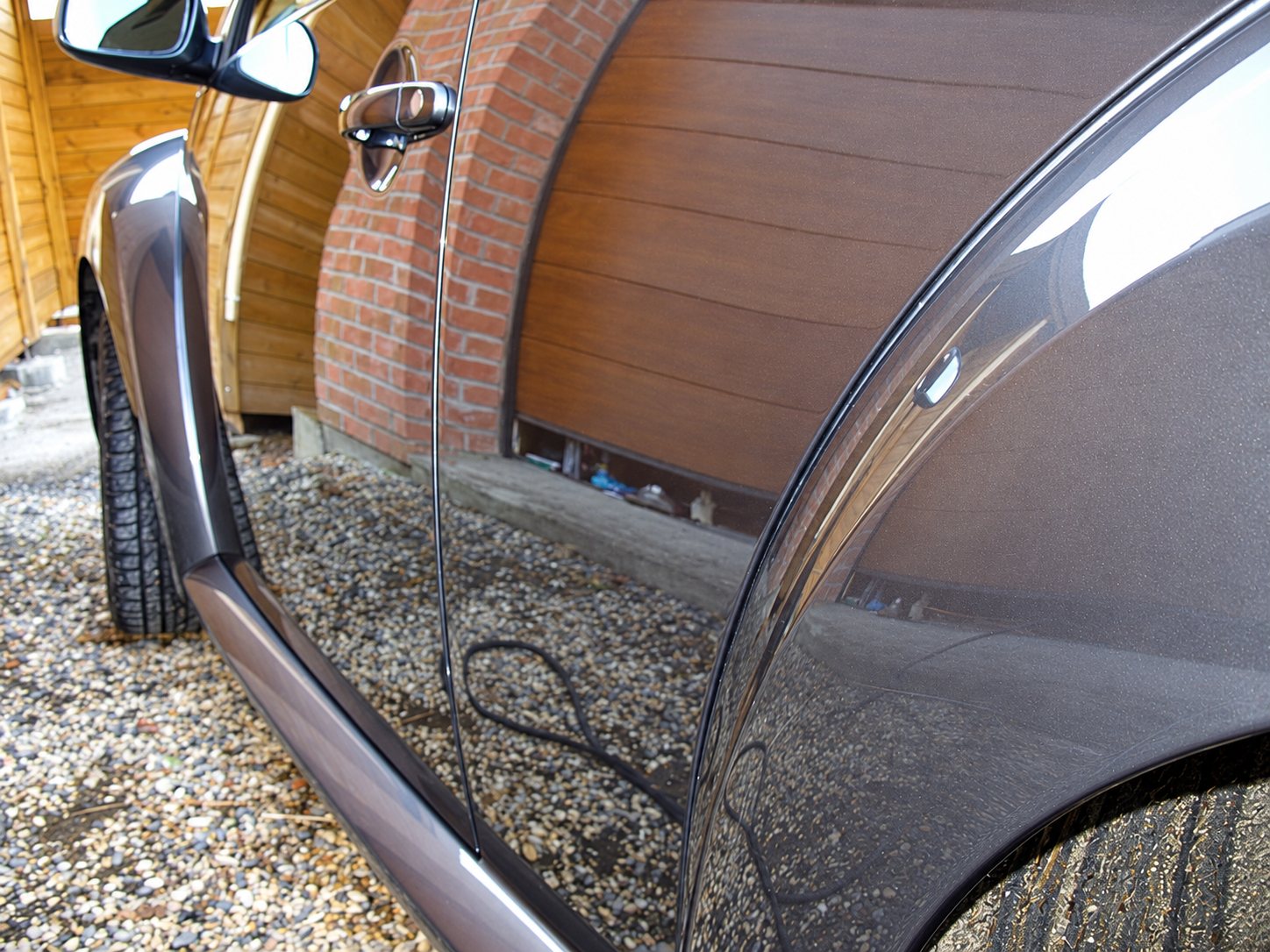Gros plan sur le côté d'une voiture grise métallisée aux reflets brillants, garée sur du gravier devant un mur de briques et une porte de garage en bois.