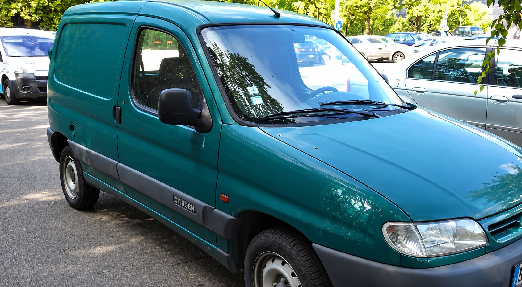 Une fourgonnette Citroën verte, un peu poussiéreuse, est garée sur un parking à côté d'autres véhicules et d'arbres.