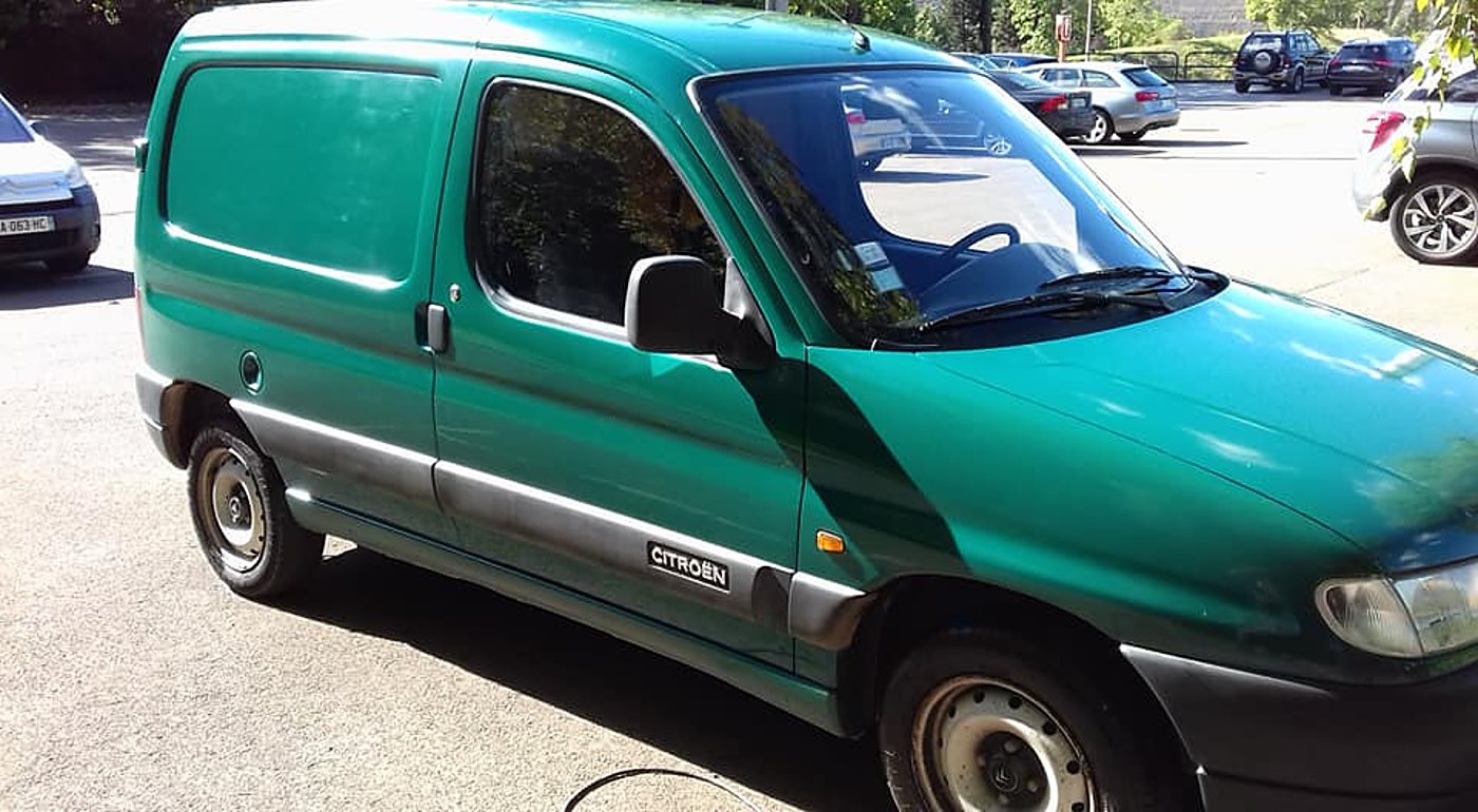 Une fourgonnette Citroën verte, vue de trois quarts avant, est garée sur un parking où stationnent d'autres véhicules.