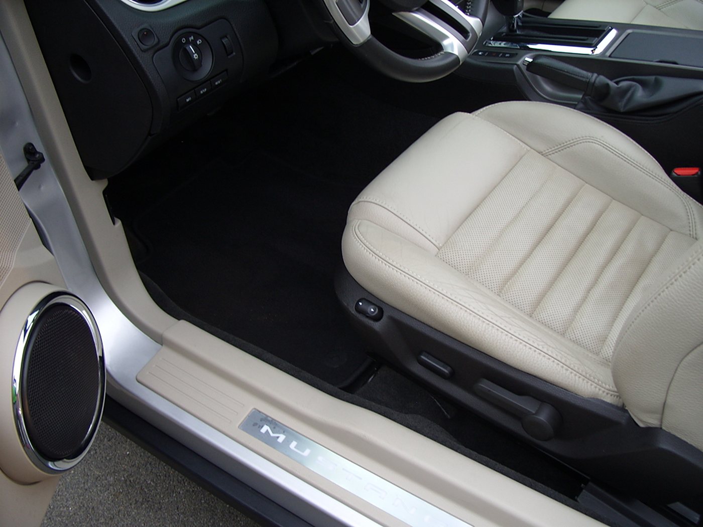 Vue de l'habitacle d'une voiture avec siège avant en cuir beige, tapis noir, haut-parleur chromé et seuil de porte métallique portant un nom en lettres partiellement visibles.
