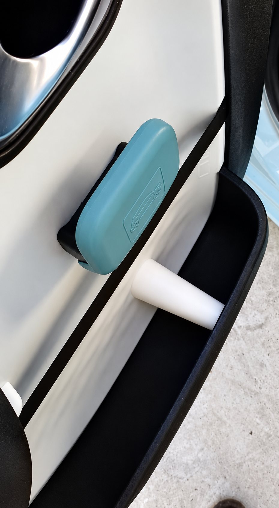 Gros plan sur l'intérieur d'une portière de voiture claire avec un petit boîtier bleu turquoise fixé et un bac de rangement noir contenant un embout blanc.