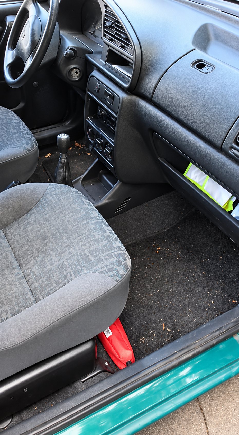 Vue de l'habitacle d'une voiture à la portière ouverte, avec un siège gris, un tapis de sol sale couvert de débris et un objet rouge sous le siège.