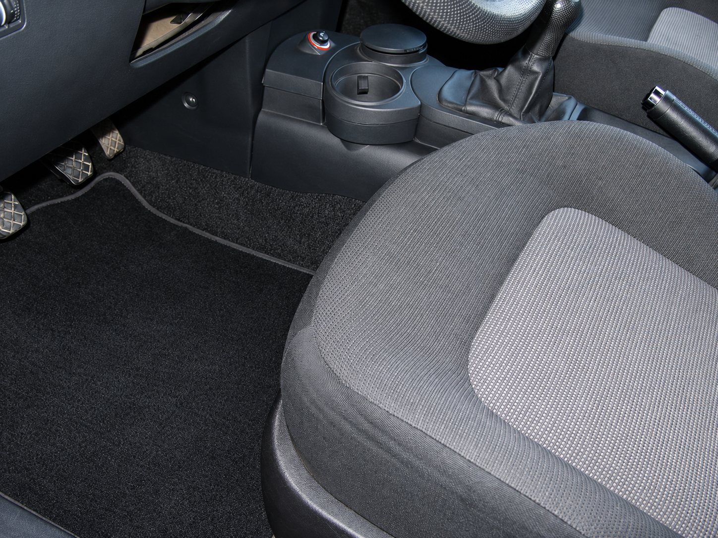 Vue de l'habitacle d'une voiture montrant un siège avant en tissu gris, un tapis de sol noir, les pédales et le porte-gobelet de la console centrale.