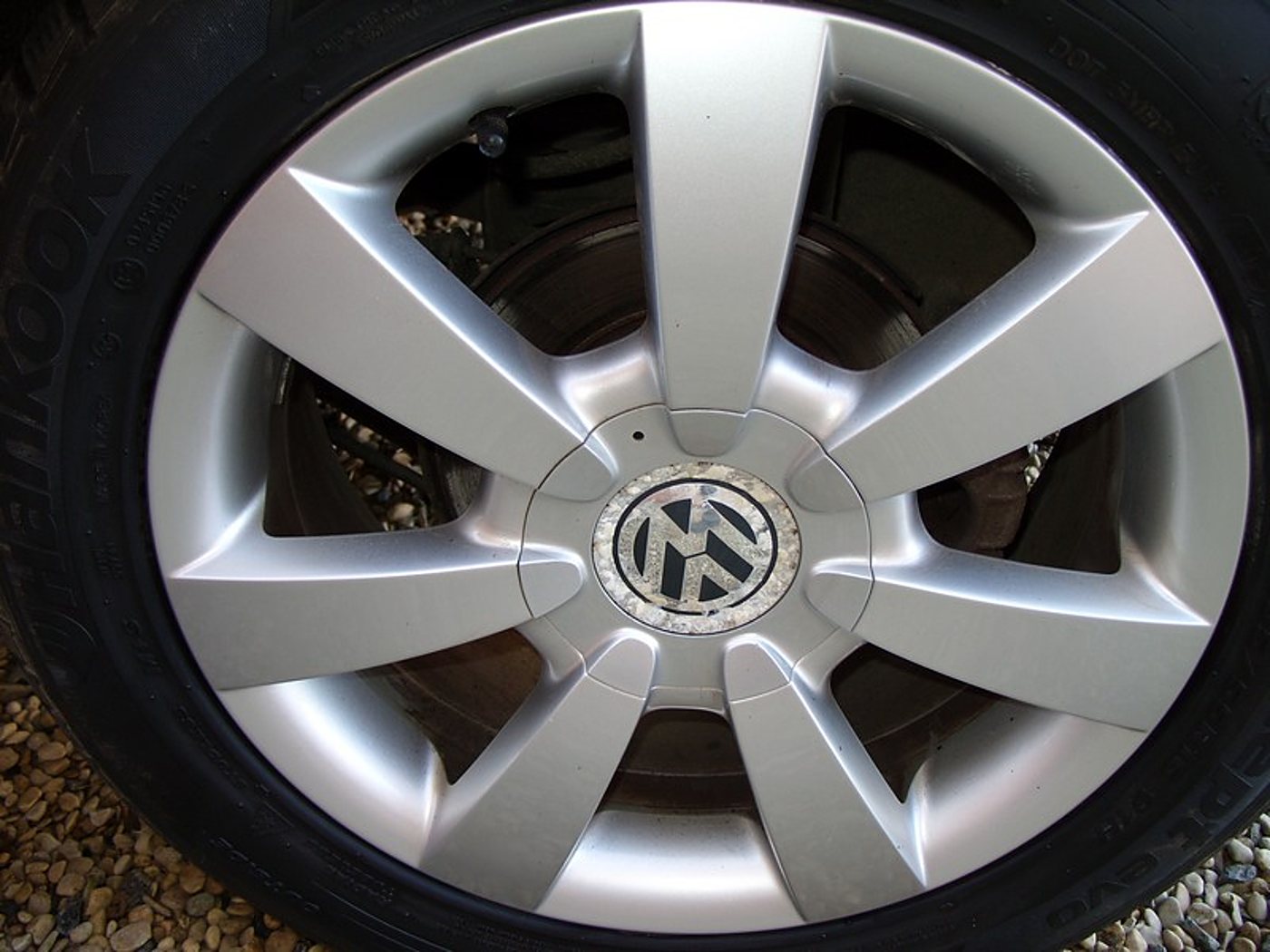 Gros plan sur une jante en alliage argenté à huit branches ornée du logo Volkswagen, montée d'un pneu noir et posée sur du gravier.