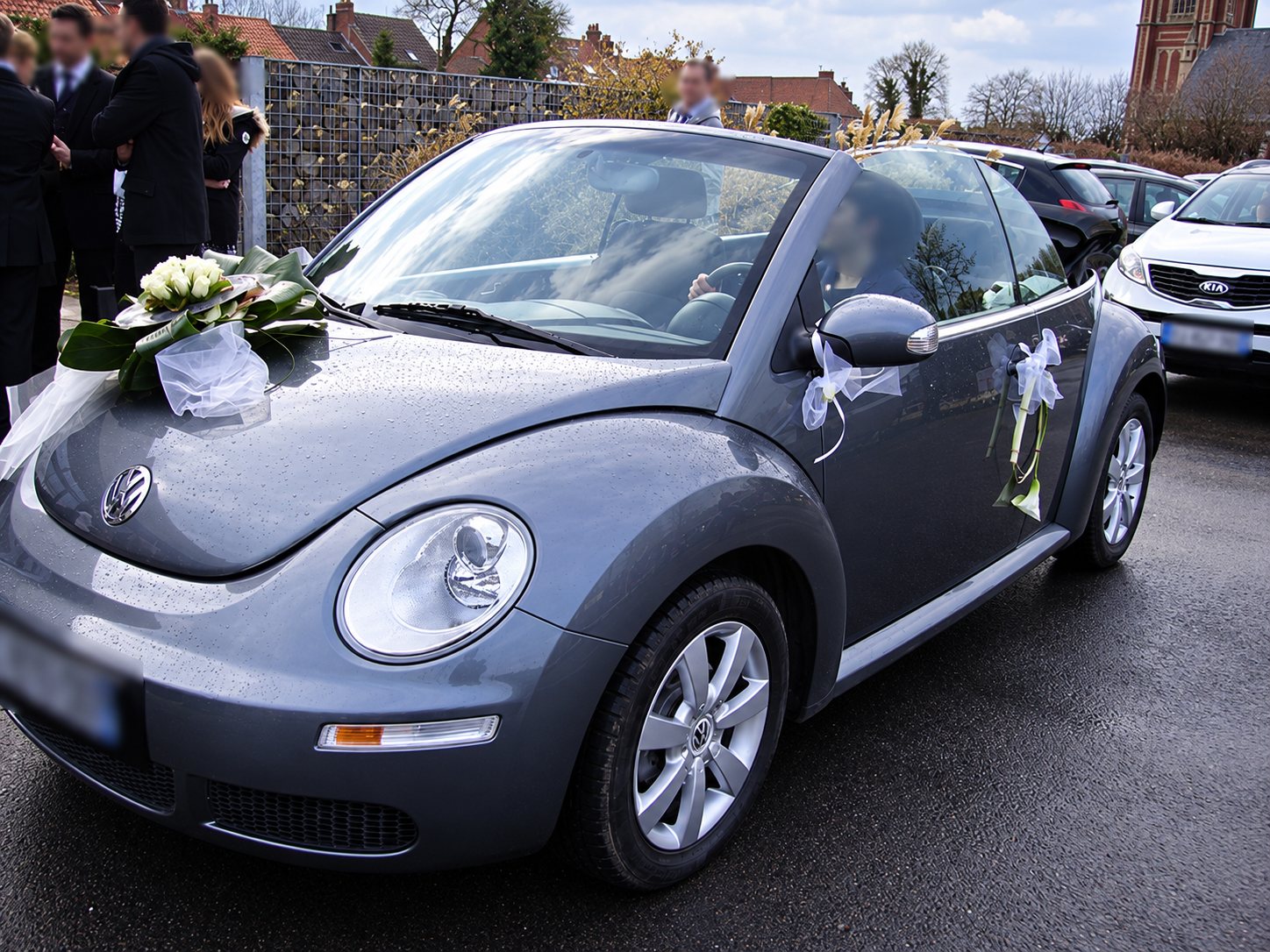 Une Volkswagen New Beetle cabriolet grise ornée d'un bouquet de fleurs blanches et de rubans de tulle pour un mariage, garée sur un parking mouillé devant des invités.