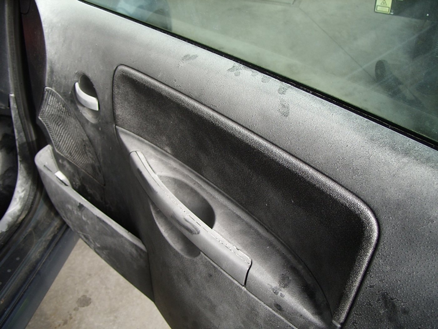 Intérieur d'une portière de voiture dont la garniture et la poignée sont recouvertes d'une épaisse couche de poussière grise, avec des traces de doigts visibles.