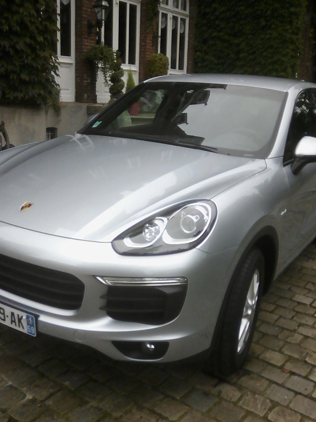 Un SUV Porsche Cayenne gris métallisé est garé sur des pavés devant une façade en briques couverte de vigne vierge.
