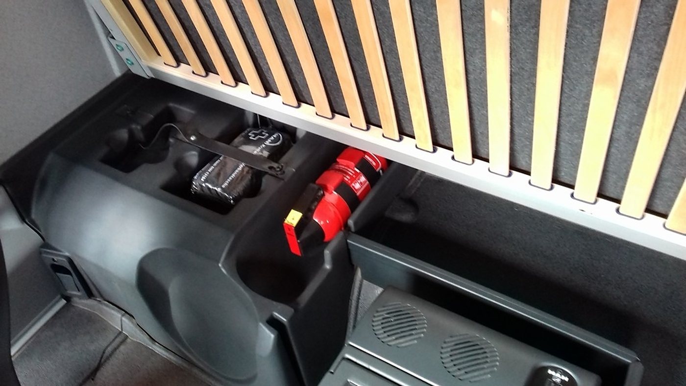 Compartiment de rangement noir sous un sommier à lattes dans un véhicule aménagé, contenant une trousse de secours et un extincteur rouge.