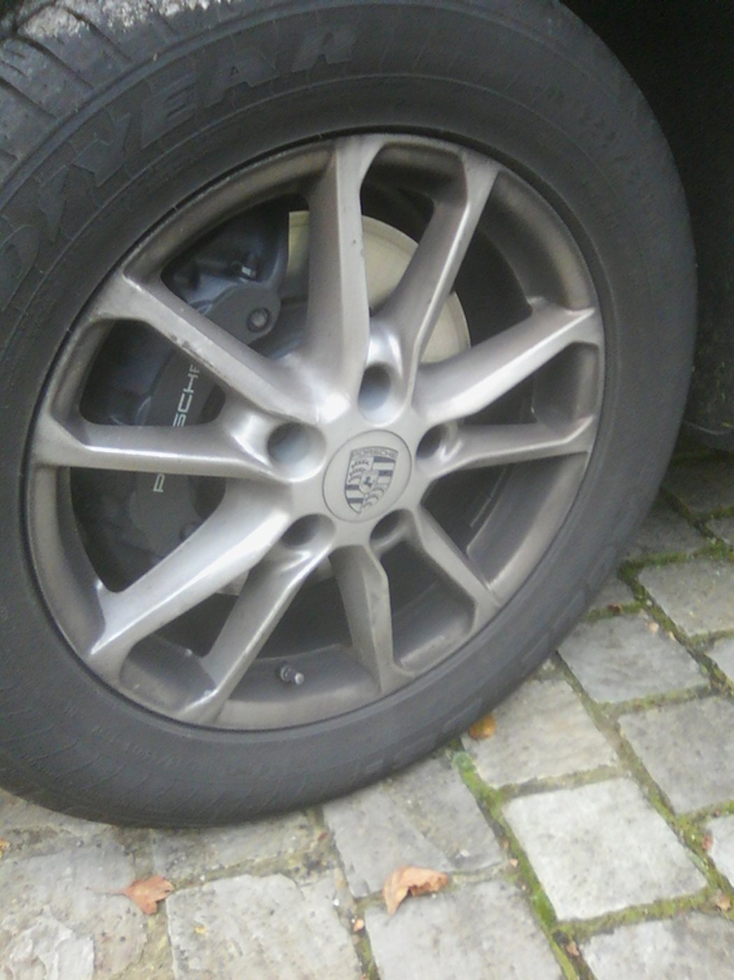 Gros plan sur une roue de Porsche avec une jante alliage grise, un étrier de frein noir et un pneu posés sur un sol pavé.
