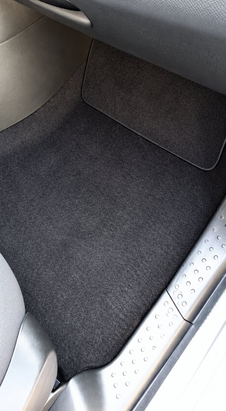 Vue du dessus d'un tapis de sol en moquette noire côté conducteur, à l'intérieur d'une voiture, près du seuil de porte métallique.