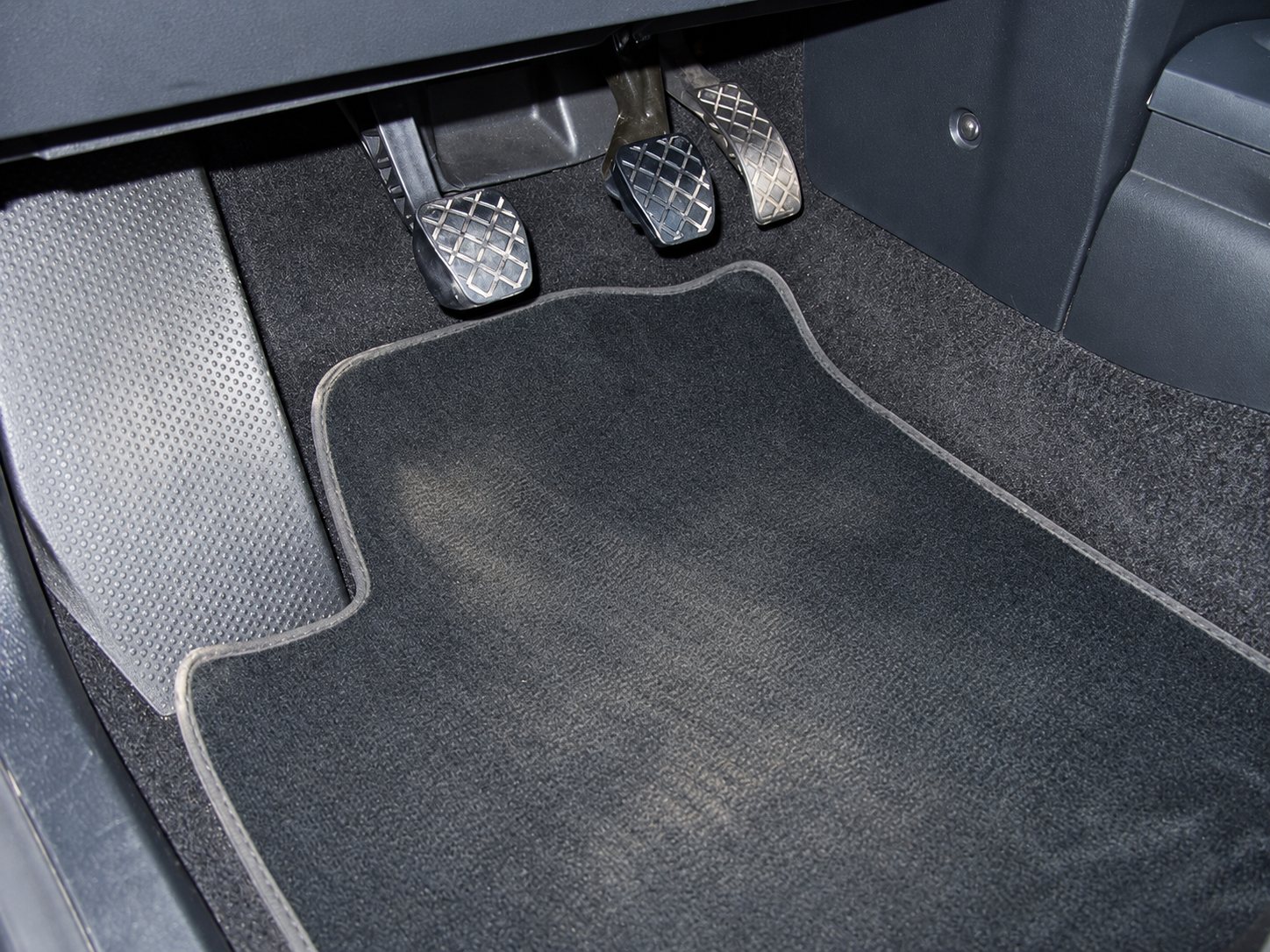 Un tapis de sol en moquette noire, légèrement poussiéreux, posé sous les pédales d'accélérateur, de frein et d'embrayage dans l'habitacle d'une voiture.