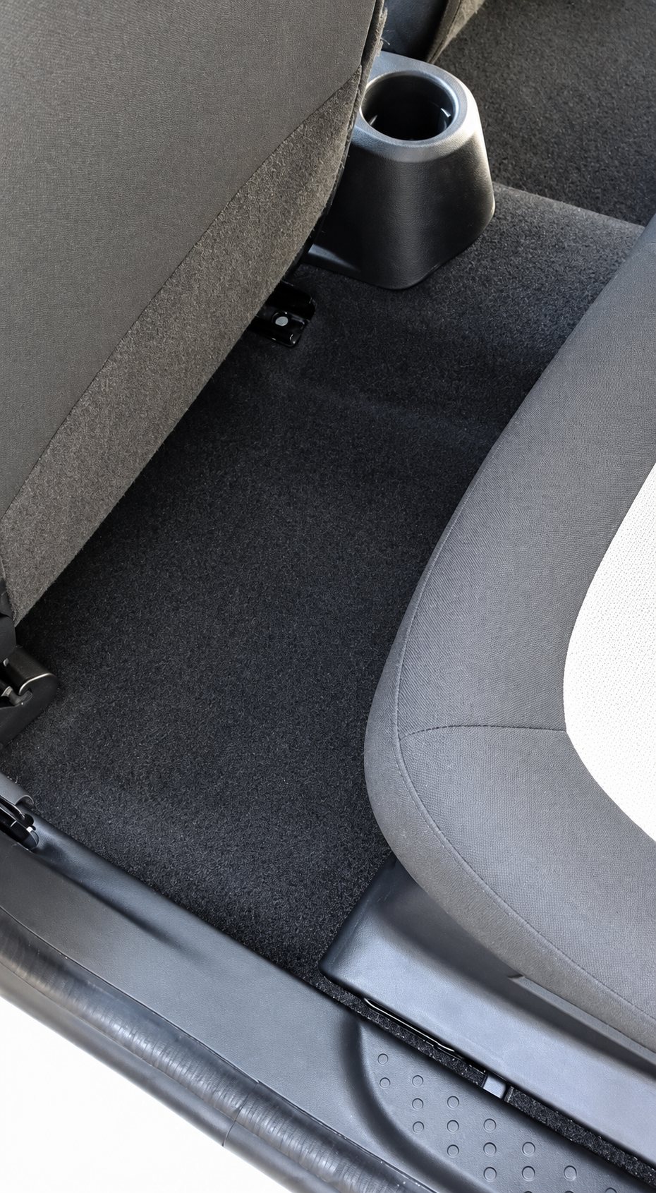 Vue de l'intérieur d'une voiture montrant un tapis de sol noir propre entre le siège avant gris et la banquette arrière, avec un porte-gobelet.