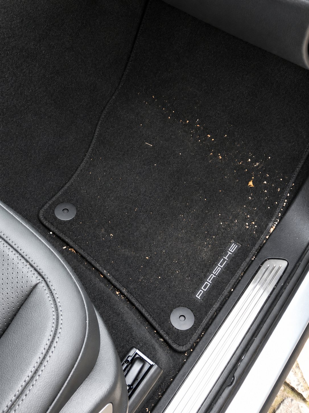 Vue du dessus d'un tapis de sol noir de marque Porsche, couvert de poussière et de débris, à côté d'un siège en cuir et d'un seuil de porte métallique.