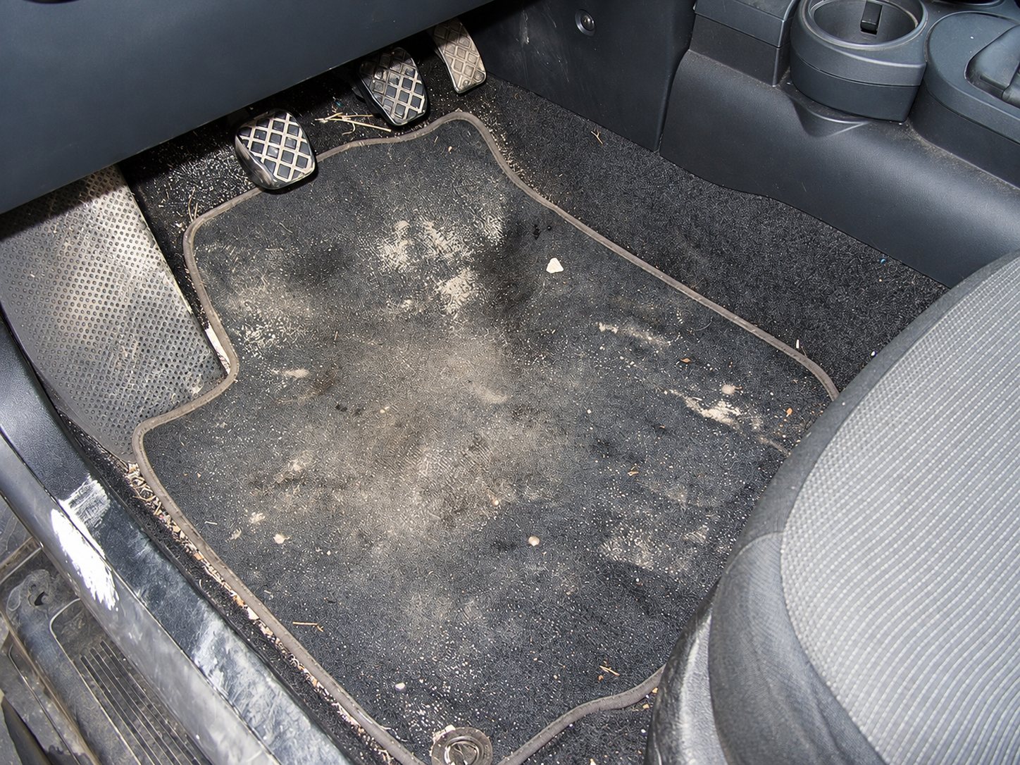 Le plancher côté conducteur d'une voiture avec un tapis noir couvert de poussière et de saletés, près des pédales et du siège.